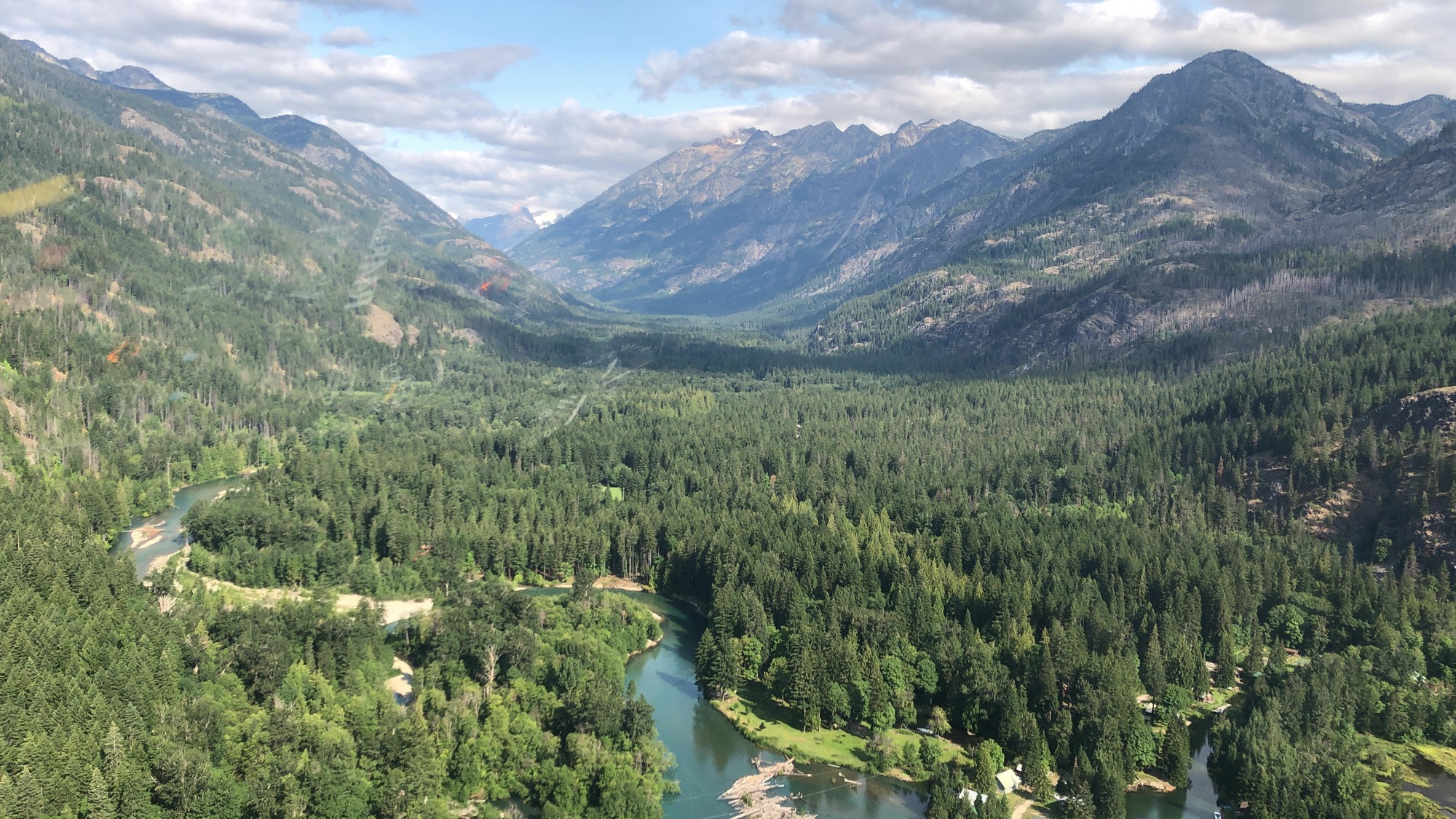 An aerial view captures a winding river flowing through a dense green forest in a vast mountain valley under a partly cloudy sky.