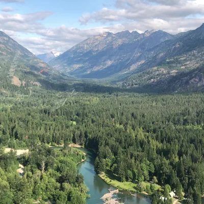 An aerial view captures a winding river flowing through a dense green forest in a vast mountain valley under a partly cloudy sky.