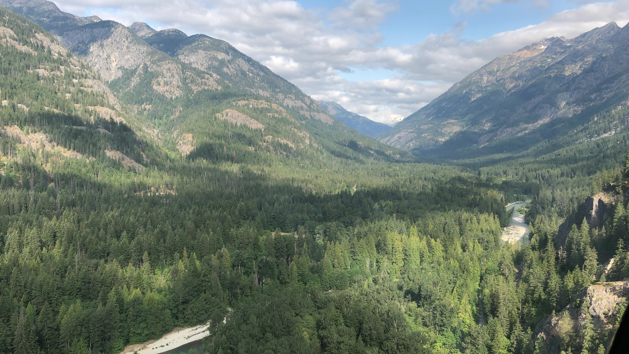 This high-angle landscape photograph captures a vast, forested mountain valley under a partly cloudy sky. A winding river is visible cutting through the dense evergreen trees below, with shadows from the clouds cast across the mountain slopes.