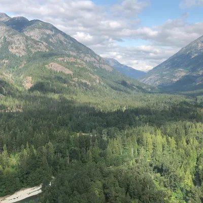 This high-angle landscape photograph captures a vast, forested mountain valley under a partly cloudy sky. A winding river is visible cutting through the dense evergreen trees below, with shadows from the clouds cast across the mountain slopes.