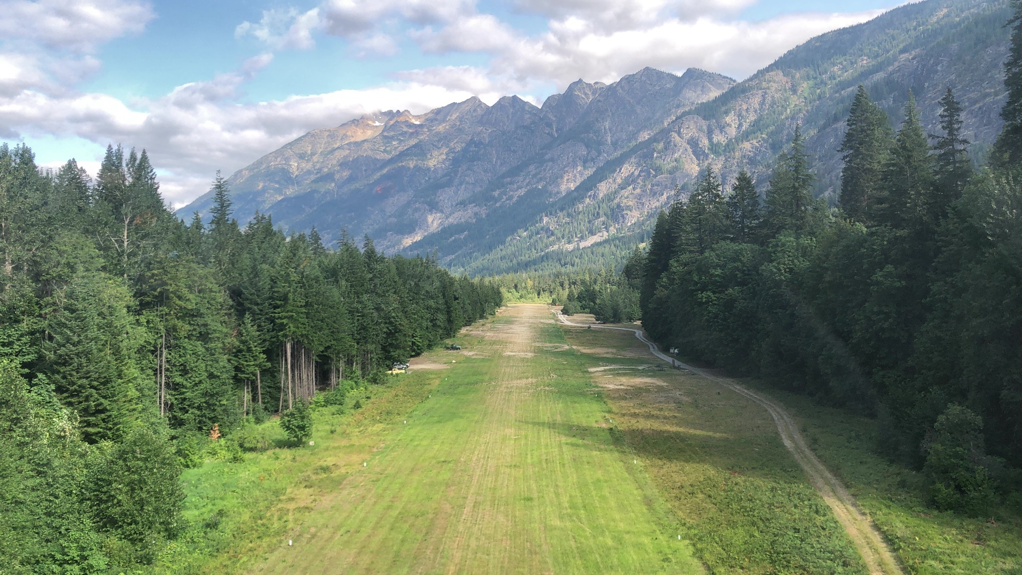 An aerial view captures a grassy airstrip stretching through a valley lined with dense evergreen forests. In the background, rugged, rocky mountains rise beneath a partly cloudy sky.