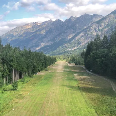 An aerial view captures a grassy airstrip stretching through a valley lined with dense evergreen forests. In the background, rugged, rocky mountains rise beneath a partly cloudy sky.