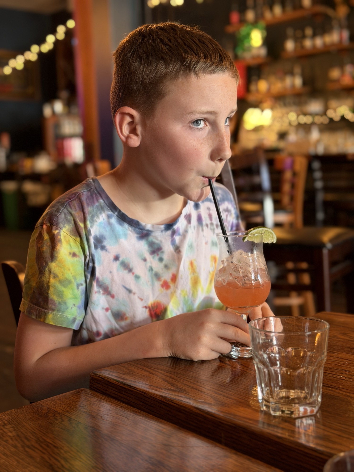 A young boy wearing a tie-dye shirt sits at a wooden table in a restaurant or bar, drinking a pink beverage with a straw and a lime wedge garnish. An empty glass sits on the table in front of him, with the dimly lit interior of the establishment visible in the background.