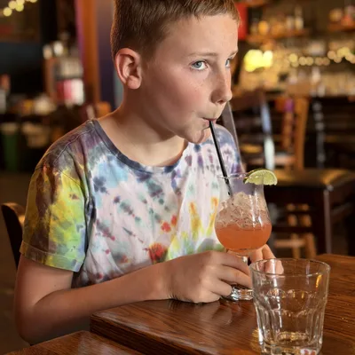 A young boy wearing a tie-dye shirt sits at a wooden table in a restaurant or bar, drinking a pink beverage with a straw and a lime wedge garnish. An empty glass sits on the table in front of him, with the dimly lit interior of the establishment visible in the background.