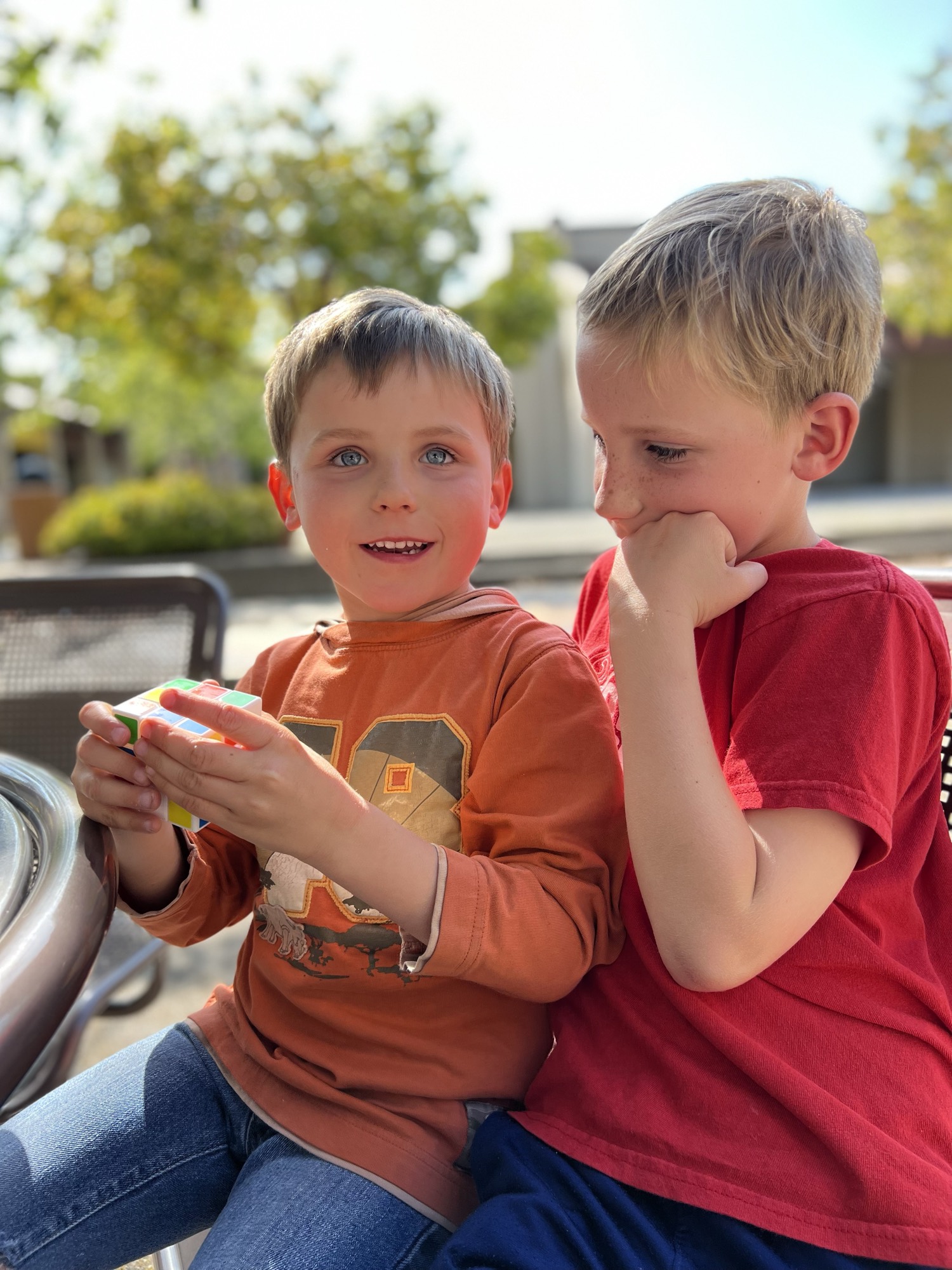 Two young boys sit together outdoors, with one holding a Rubik's cube and looking toward the camera while the other watches him with his hand on his chin. They are seated at a table in a sunny, paved outdoor area with trees and buildings blurred in the background.