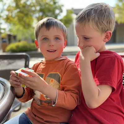 Two young boys sit together outdoors, with one holding a Rubik's cube and looking toward the camera while the other watches him with his hand on his chin. They are seated at a table in a sunny, paved outdoor area with trees and buildings blurred in the background.