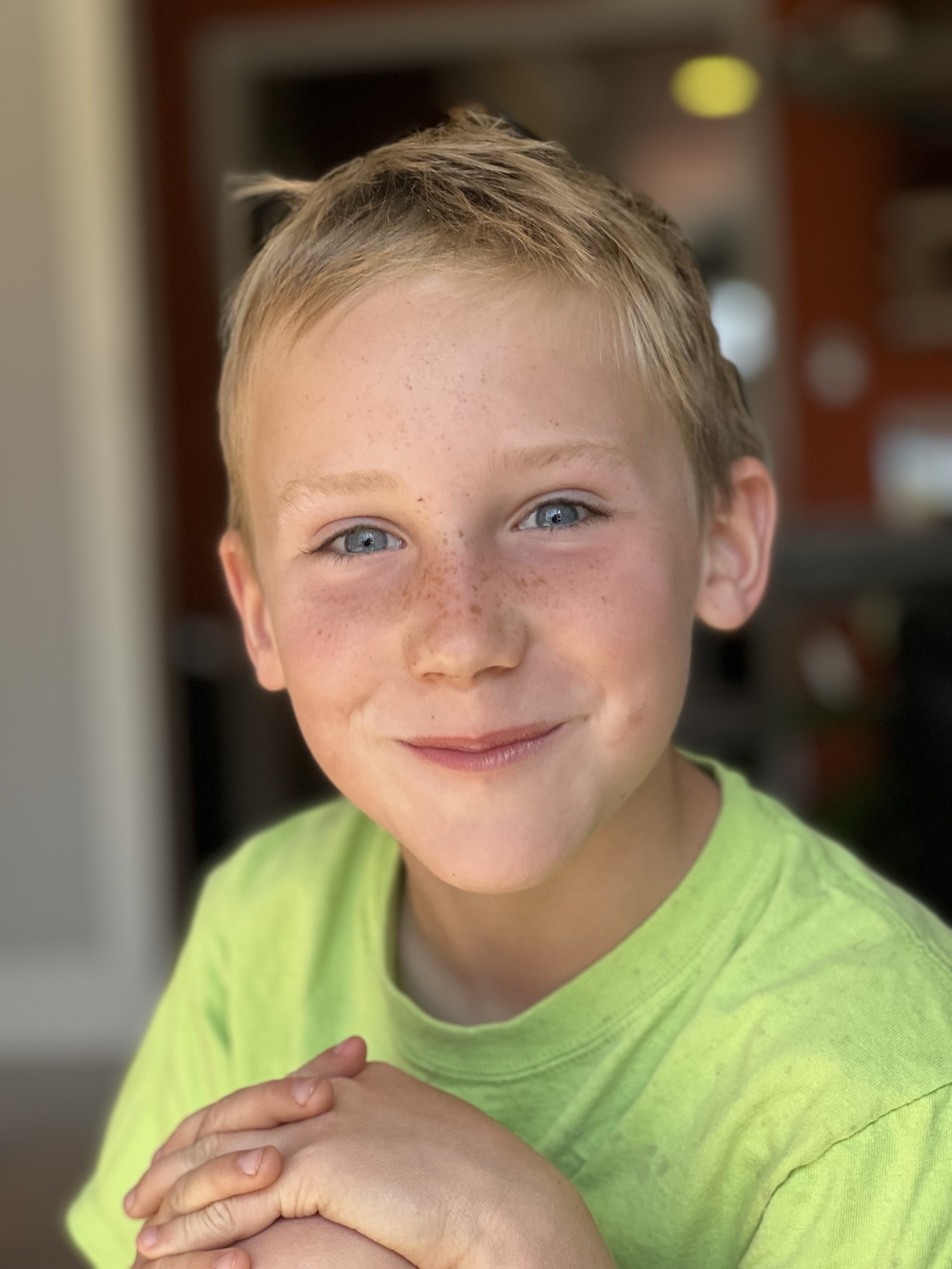 A close-up portrait of a young boy with blonde hair, blue eyes, and freckles smiling directly at the camera. He is wearing a bright green t-shirt and has his hands clasped in front of him, set against a softly blurred indoor background.