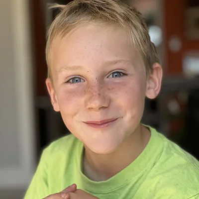 A close-up portrait of a young boy with blonde hair, blue eyes, and freckles smiling directly at the camera. He is wearing a bright green t-shirt and has his hands clasped in front of him, set against a softly blurred indoor background.