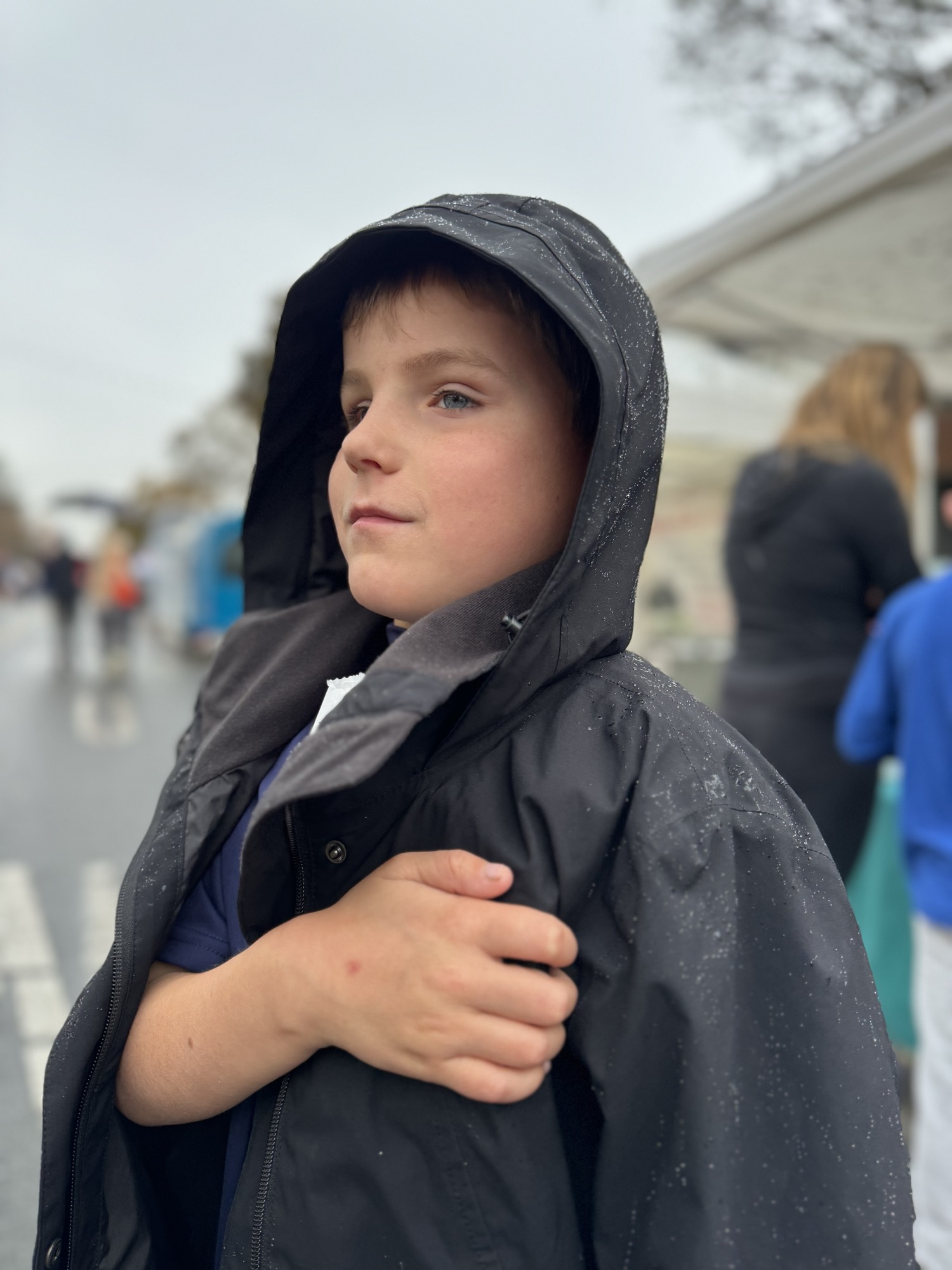 A young boy wearing a black hooded jacket with water droplets on it stands outdoors on a rainy day, looking off to the side. The background is softly blurred, showing other people and what appears to be an outdoor street or market setting.