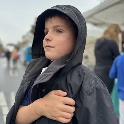 A young boy wearing a black hooded jacket with water droplets on it stands outdoors on a rainy day, looking off to the side. The background is softly blurred, showing other people and what appears to be an outdoor street or market setting.