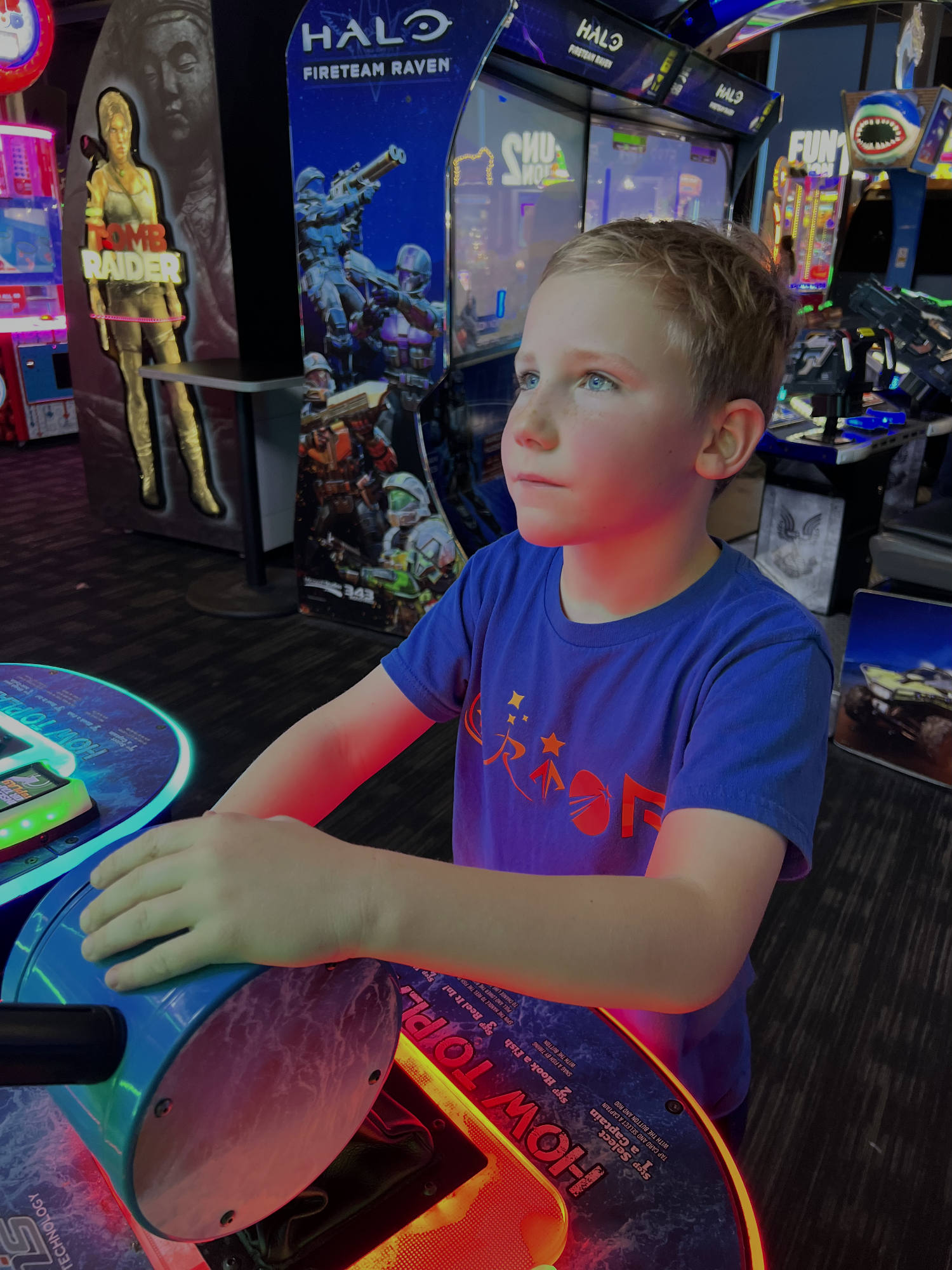 A young boy in a blue t-shirt stands at an arcade game, holding a large blue controller while looking up at the screen. The background is filled with various brightly lit arcade cabinets, including "Halo: Fireteam Raven" and "Tomb Raider."