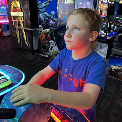 A young boy in a blue t-shirt stands at an arcade game, holding a large blue controller while looking up at the screen. The background is filled with various brightly lit arcade cabinets, including "Halo: Fireteam Raven" and "Tomb Raider."