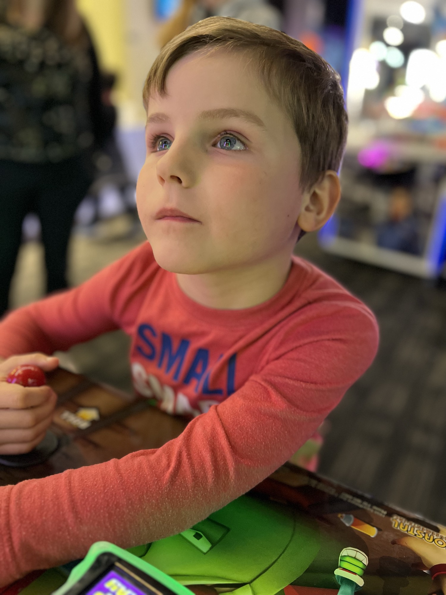 A young boy in a red long-sleeved shirt looks upward while holding a joystick at an arcade game. The background is softly blurred, showing other arcade machines and lights.
