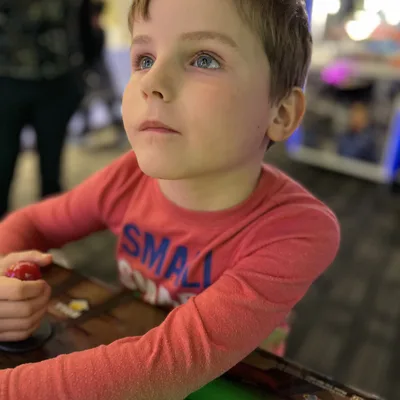 A young boy in a red long-sleeved shirt looks upward while holding a joystick at an arcade game. The background is softly blurred, showing other arcade machines and lights.