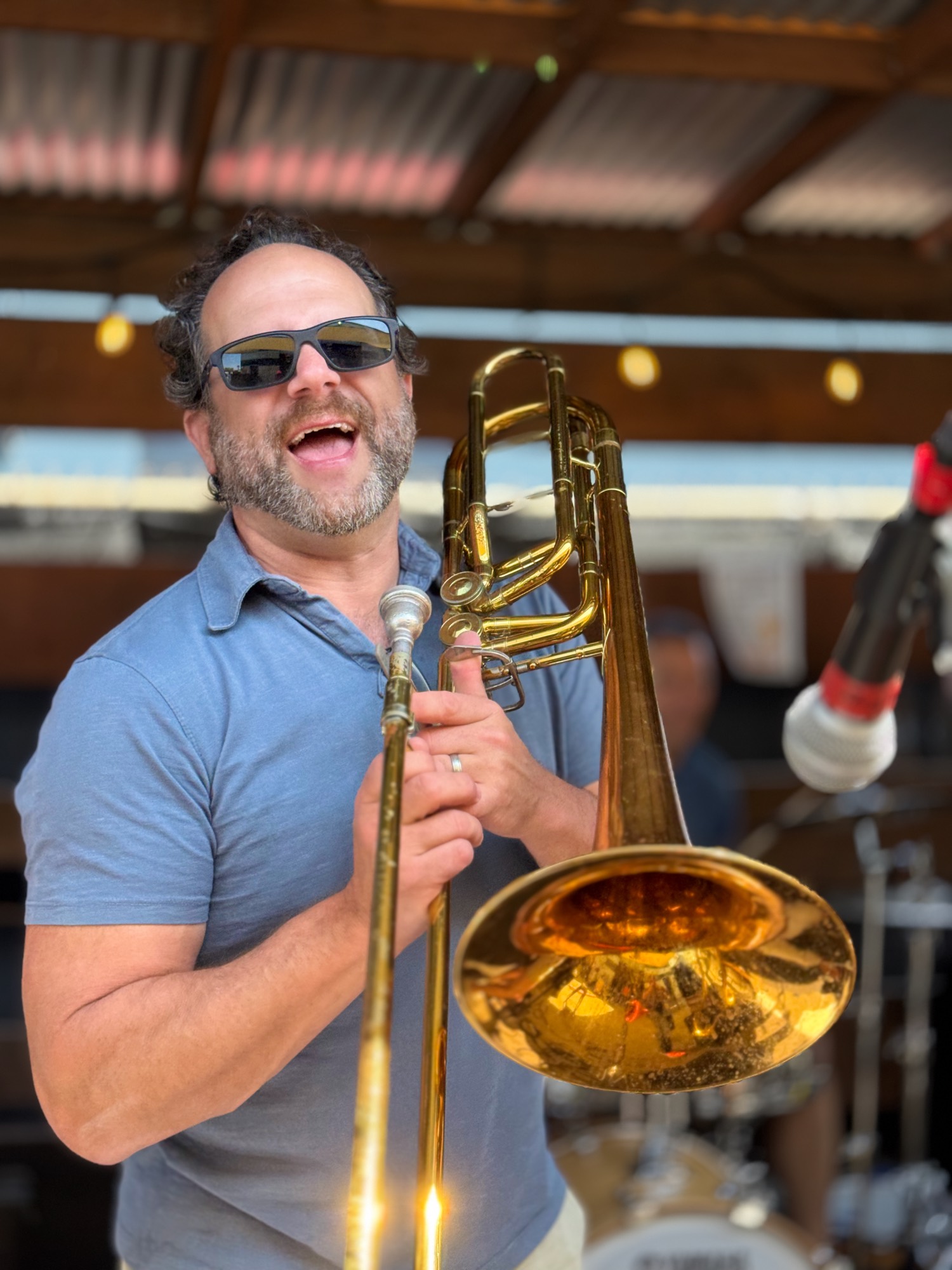 A smiling man wearing sunglasses and a blue polo shirt holds a brass trombone on an outdoor stage. He appears to be performing, with a microphone positioned nearby and a drum kit visible in the blurred background.