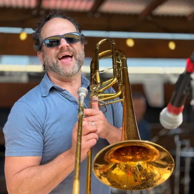 A smiling man wearing sunglasses and a blue polo shirt holds a brass trombone on an outdoor stage. He appears to be performing, with a microphone positioned nearby and a drum kit visible in the blurred background.