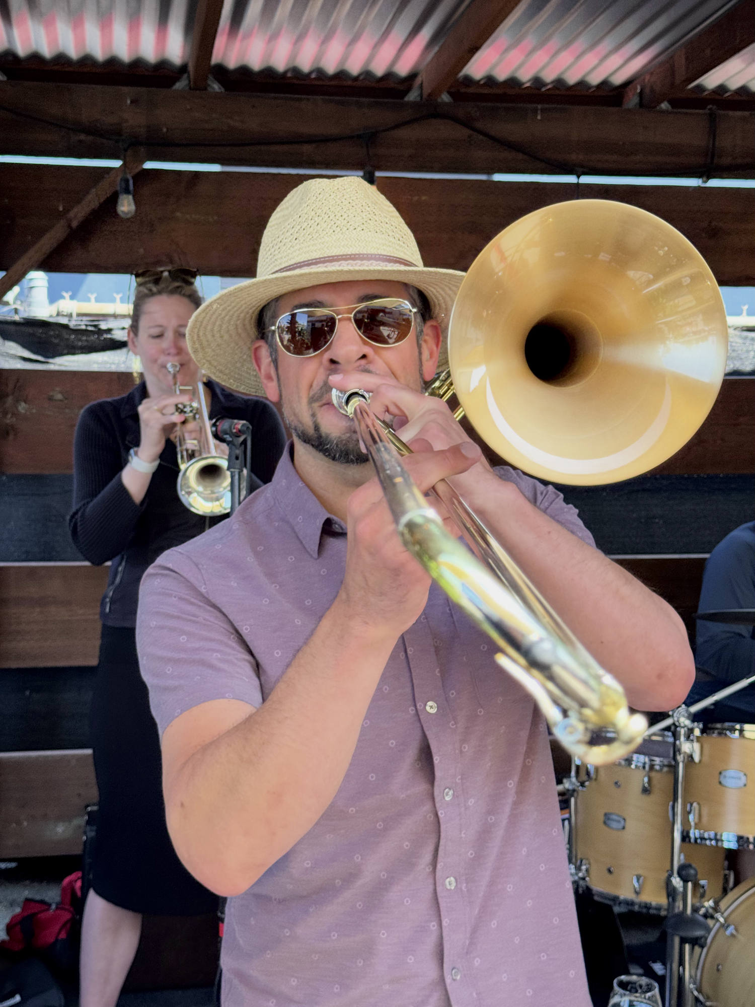 A man wearing a straw hat and sunglasses plays a trombone in the foreground, while a woman plays a trumpet behind him on an outdoor covered stage. A drum set is partially visible to the right of the trombone player.