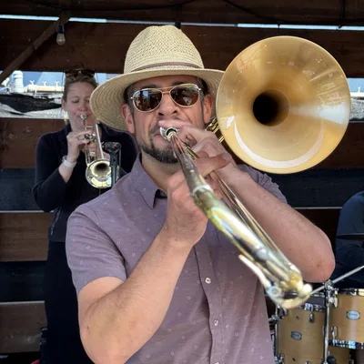 A man wearing a straw hat and sunglasses plays a trombone in the foreground, while a woman plays a trumpet behind him on an outdoor covered stage. A drum set is partially visible to the right of the trombone player.