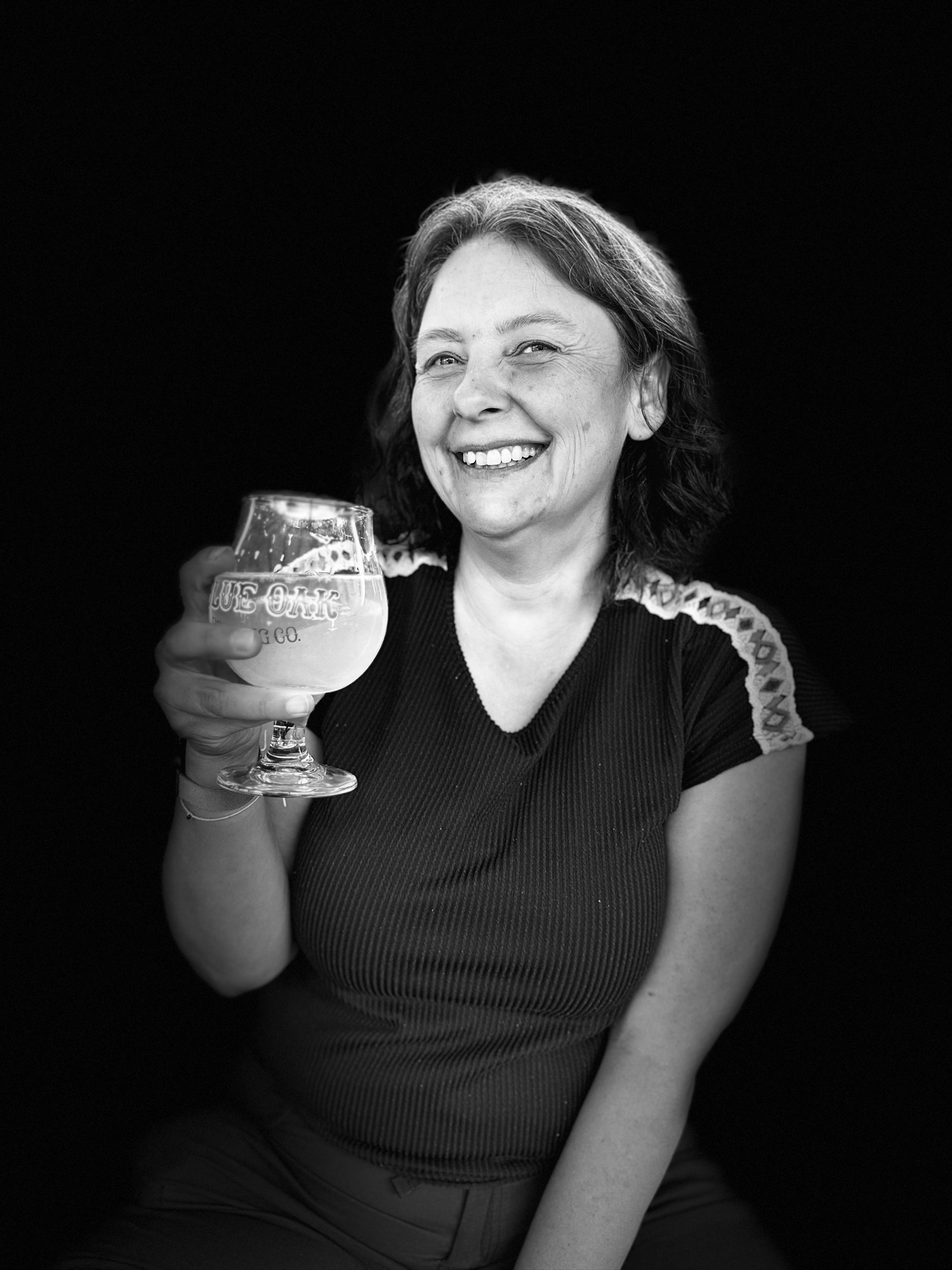 A black and white portrait of a smiling woman holding up a glass of beer against a solid black background.