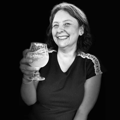 A black and white portrait of a smiling woman holding up a glass of beer against a solid black background.