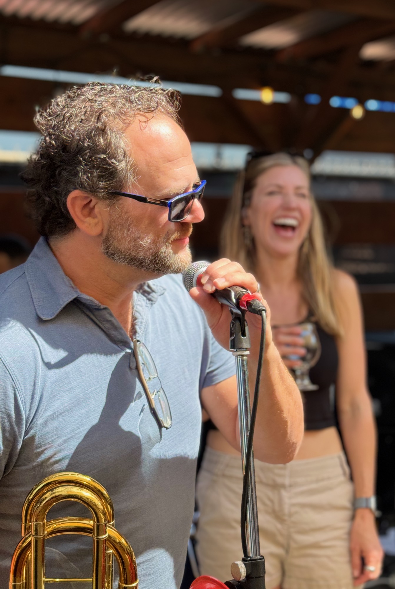 A man wearing sunglasses speaks or sings into a microphone while holding a brass instrument, with a laughing woman holding a drink visible in the background. The outdoor setting appears to be a patio or stage area with a wooden roof structure overhead.