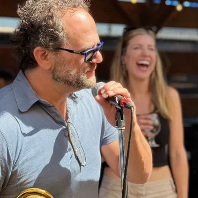 A man wearing sunglasses speaks or sings into a microphone while holding a brass instrument, with a laughing woman holding a drink visible in the background. The outdoor setting appears to be a patio or stage area with a wooden roof structure overhead.