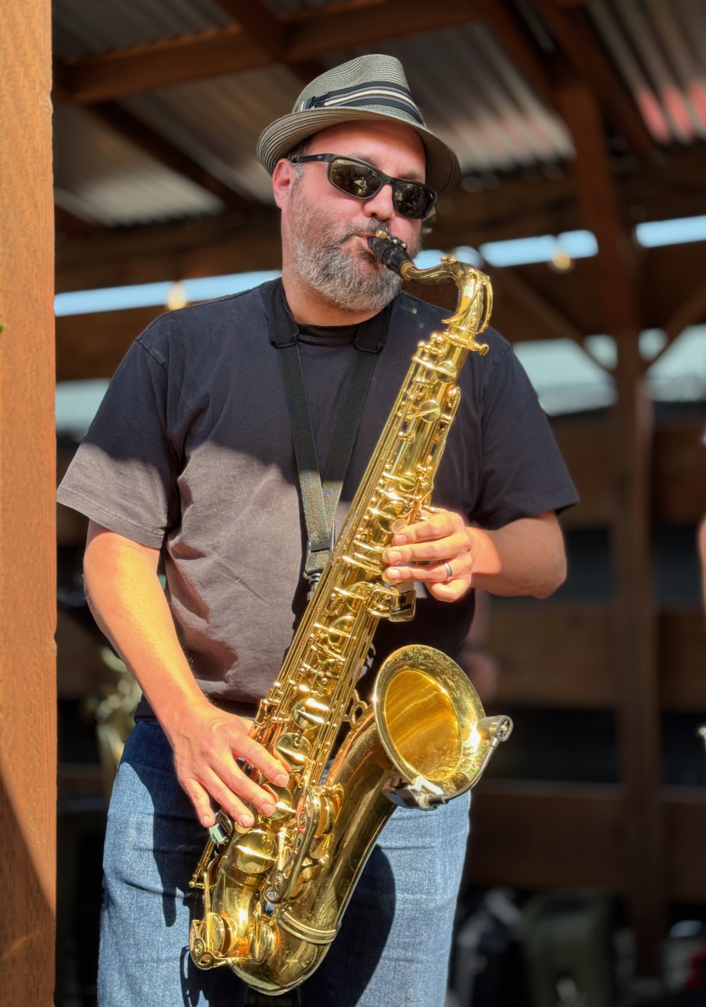 A man wearing a hat, sunglasses, and a black t-shirt plays a brass saxophone outdoors. He is standing under a wooden structure with a corrugated metal roof.