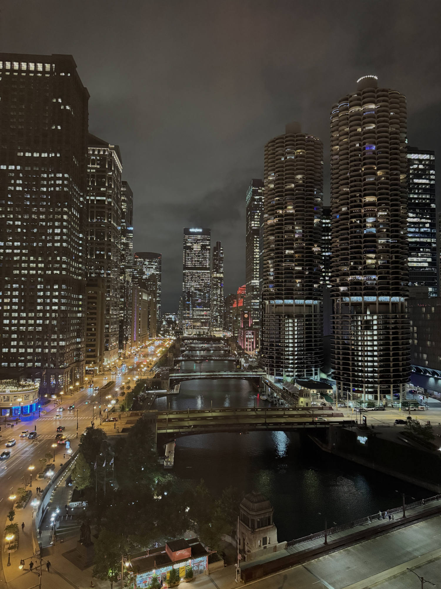 An elevated, nighttime view of the Chicago River flowing through downtown Chicago, flanked by illuminated skyscrapers, including the Marina City towers on the right. Streetlights and vehicle headlights light up Wacker Drive on the left, while several bridges span the dark water under a cloudy night sky.