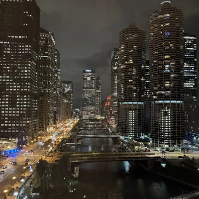 An elevated, nighttime view of the Chicago River flowing through downtown Chicago, flanked by illuminated skyscrapers, including the Marina City towers on the right. Streetlights and vehicle headlights light up Wacker Drive on the left, while several bridges span the dark water under a cloudy night sky.