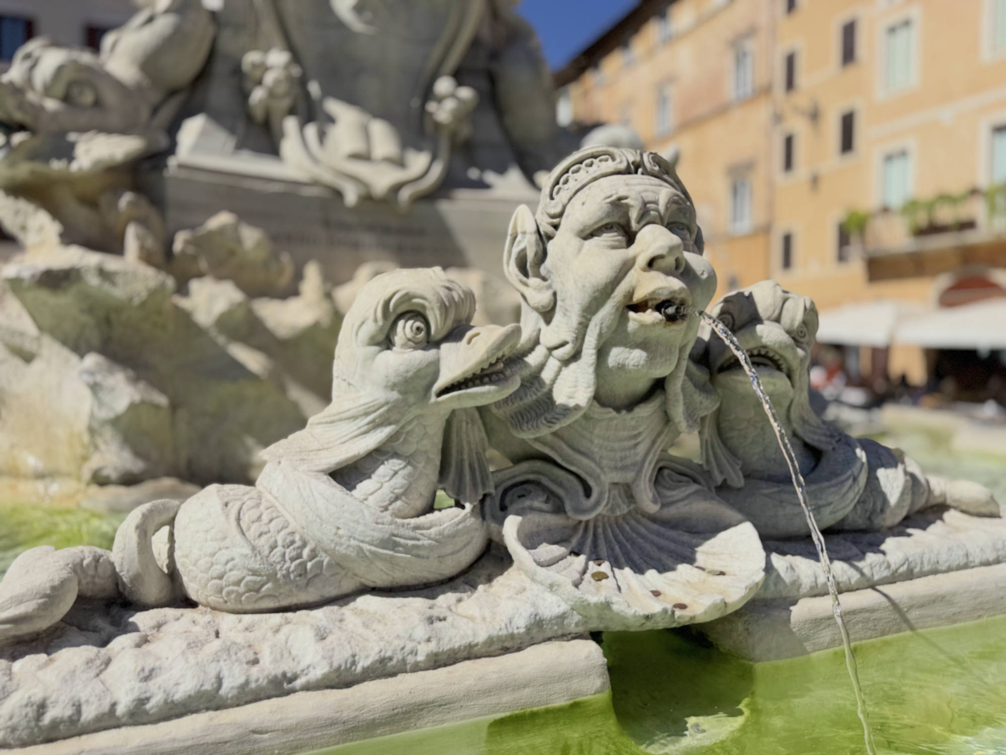 A close-up shot captures a stone fountain sculpture featuring a central human-like face flanked by two stylized fish, with water spouting from the figure's mouth into the basin below. The outdoor setting includes blurred buildings in the background under a clear blue sky.