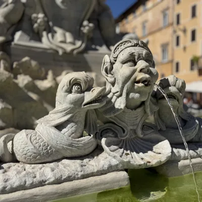 A close-up shot captures a stone fountain sculpture featuring a central human-like face flanked by two stylized fish, with water spouting from the figure's mouth into the basin below. The outdoor setting includes blurred buildings in the background under a clear blue sky.
