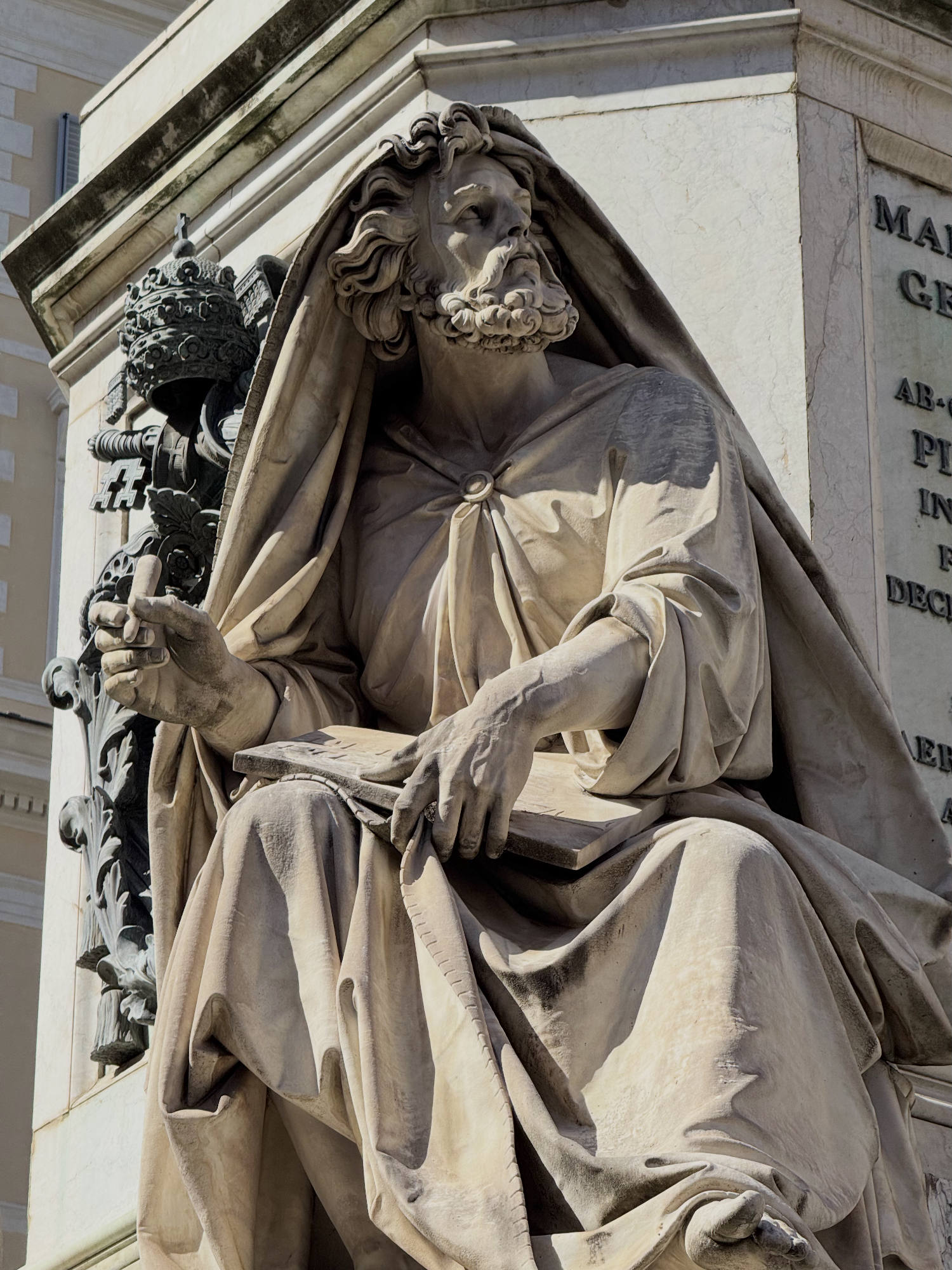 This close-up shot features a marble statue of a bearded, draped figure holding a tablet and looking upward, situated outdoors against a stone monument. To the left of the figure, a dark metal papal tiara and keys emblem is mounted on the stone structure.