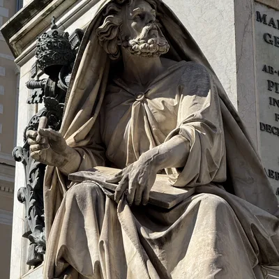 This close-up shot features a marble statue of a bearded, draped figure holding a tablet and looking upward, situated outdoors against a stone monument. To the left of the figure, a dark metal papal tiara and keys emblem is mounted on the stone structure.