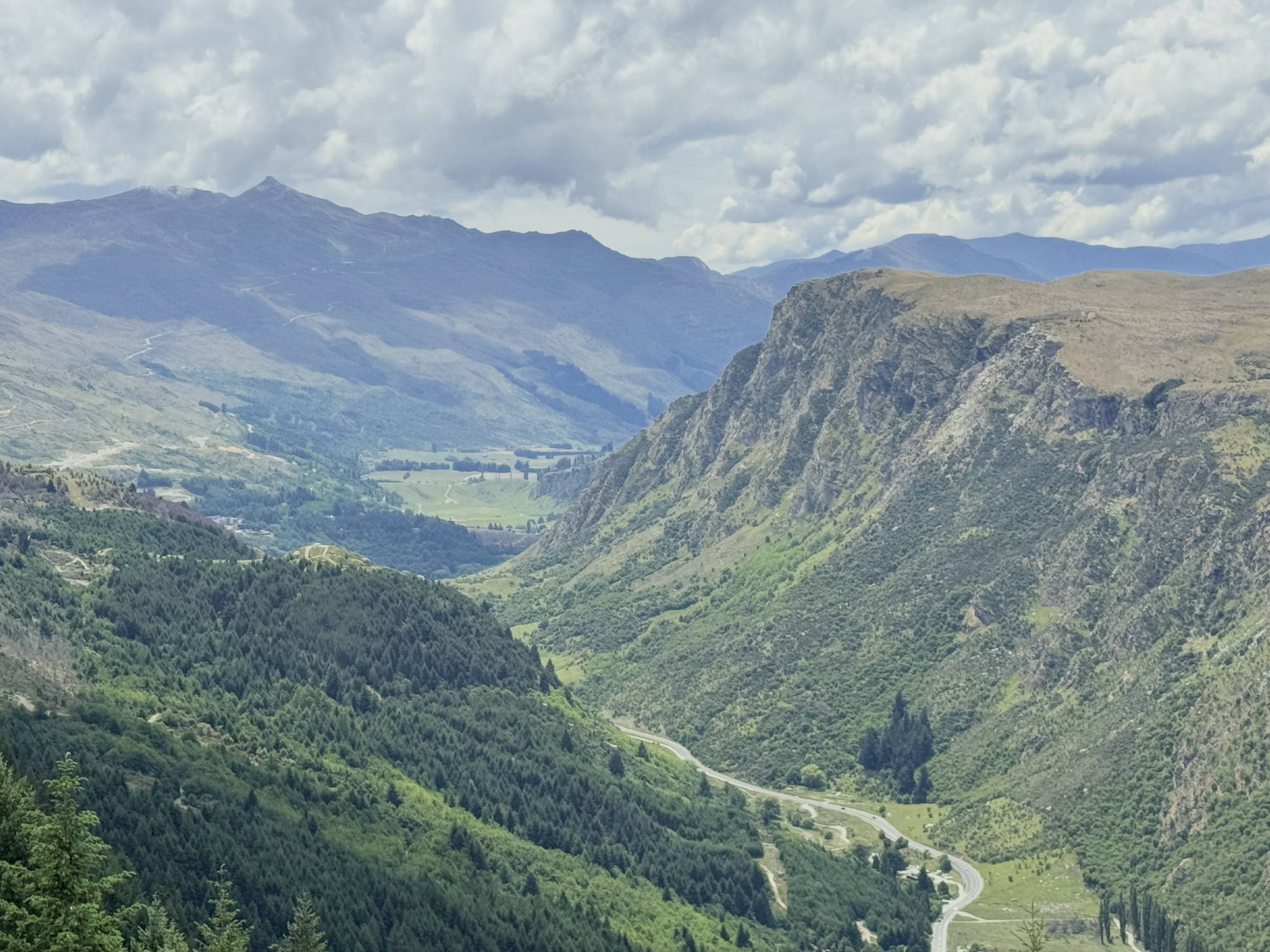 This high-angle landscape photograph captures a vast mountain valley with steep, green slopes and a winding road visible on the valley floor. In the background, layered mountain ranges stretch beneath a cloudy sky.