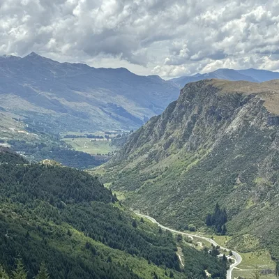 This high-angle landscape photograph captures a vast mountain valley with steep, green slopes and a winding road visible on the valley floor. In the background, layered mountain ranges stretch beneath a cloudy sky.