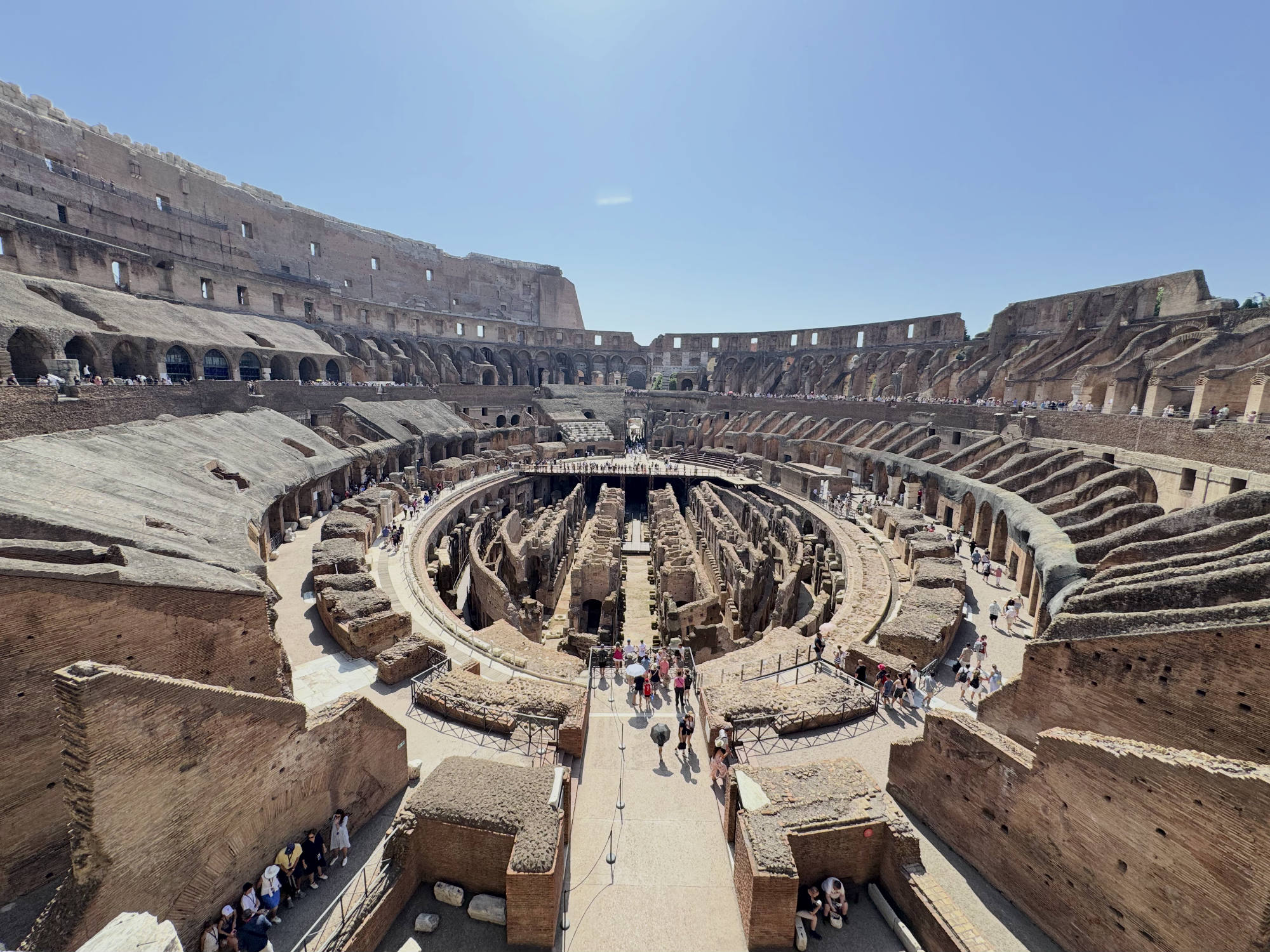 This high-angle shot captures tourists exploring the interior of the Colosseum in Rome, Italy, under a clear blue sky. The image highlights the exposed underground tunnels of the hypogeum, surrounded by the tiered stone seating areas of the ancient amphitheater.