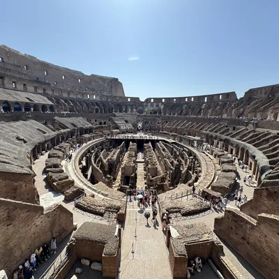 This high-angle shot captures tourists exploring the interior of the Colosseum in Rome, Italy, under a clear blue sky. The image highlights the exposed underground tunnels of the hypogeum, surrounded by the tiered stone seating areas of the ancient amphitheater.
