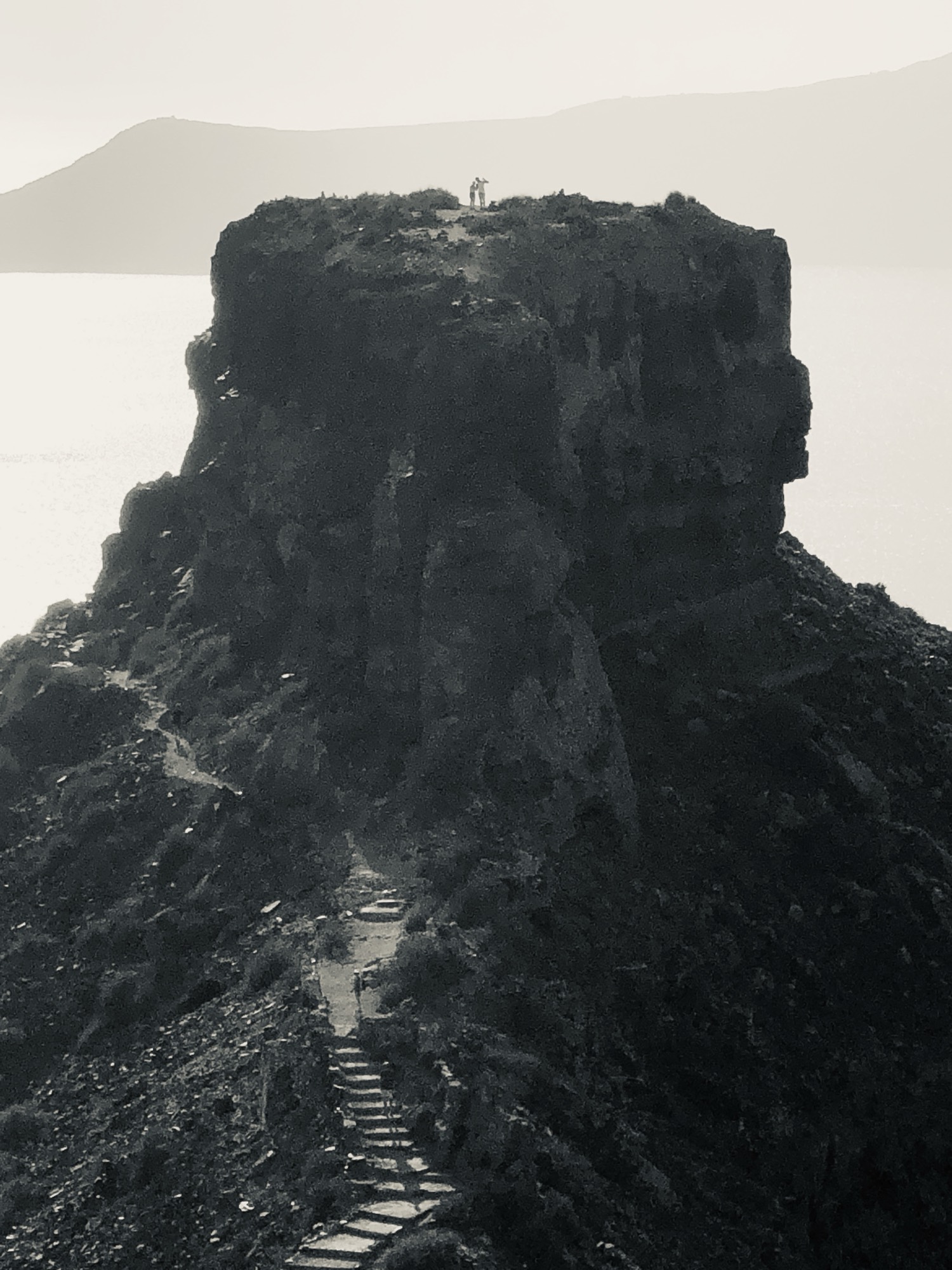 A black-and-white photograph captures a massive, dark rock formation rising above a body of water, with a narrow, winding path of stone steps leading up toward it. At the very top of the peak, two small figures of people stand looking out over the misty landscape.