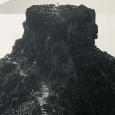 A black-and-white photograph captures a massive, dark rock formation rising above a body of water, with a narrow, winding path of stone steps leading up toward it. At the very top of the peak, two small figures of people stand looking out over the misty landscape.