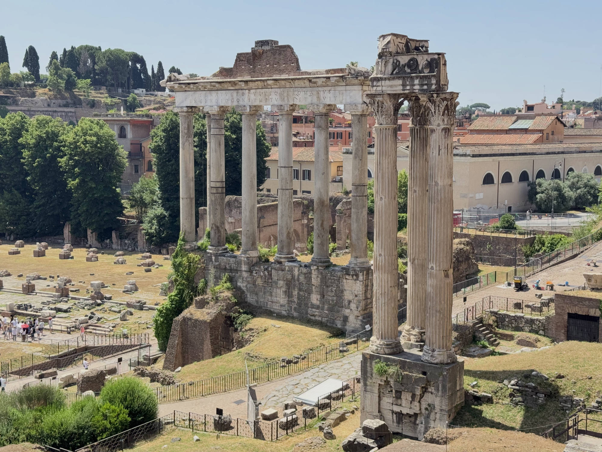 An elevated, wide shot captures the Temple of Saturn, featuring its standing ionic columns and architrave, situated within the Roman Forum in Rome, Italy. Tourists walk along the pathways below the ancient ruins on a clear, sunny day.