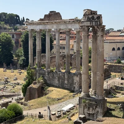 An elevated, wide shot captures the Temple of Saturn, featuring its standing ionic columns and architrave, situated within the Roman Forum in Rome, Italy. Tourists walk along the pathways below the ancient ruins on a clear, sunny day.