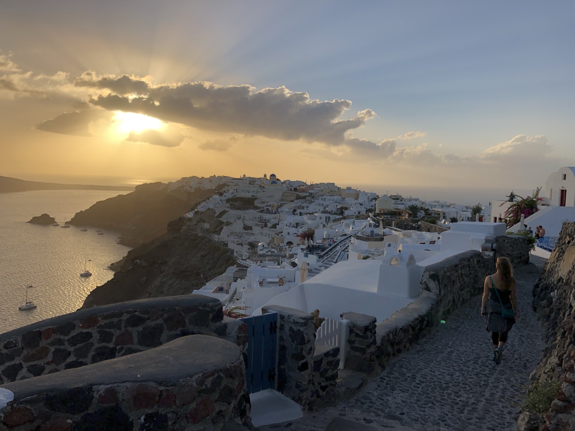 A person walks along a stone path overlooking a hillside village of white buildings in Santorini, Greece, during a golden sunset. The sun shines through clouds over the sea, casting a warm glow across the coastal landscape.