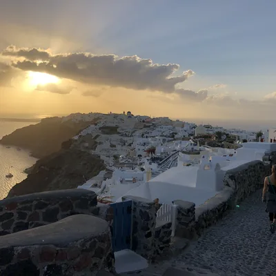 A person walks along a stone path overlooking a hillside village of white buildings in Santorini, Greece, during a golden sunset. The sun shines through clouds over the sea, casting a warm glow across the coastal landscape.