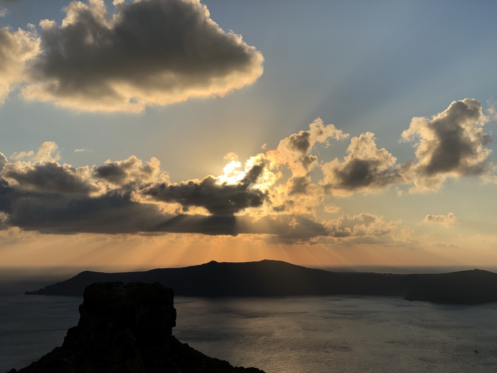 A scenic view of a sunset over a body of water, with sunbeams filtering through clouds above a silhouetted island and a rocky outcrop in the foreground.
