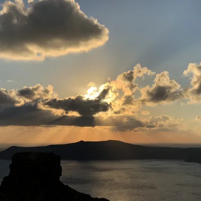 A scenic view of a sunset over a body of water, with sunbeams filtering through clouds above a silhouetted island and a rocky outcrop in the foreground.