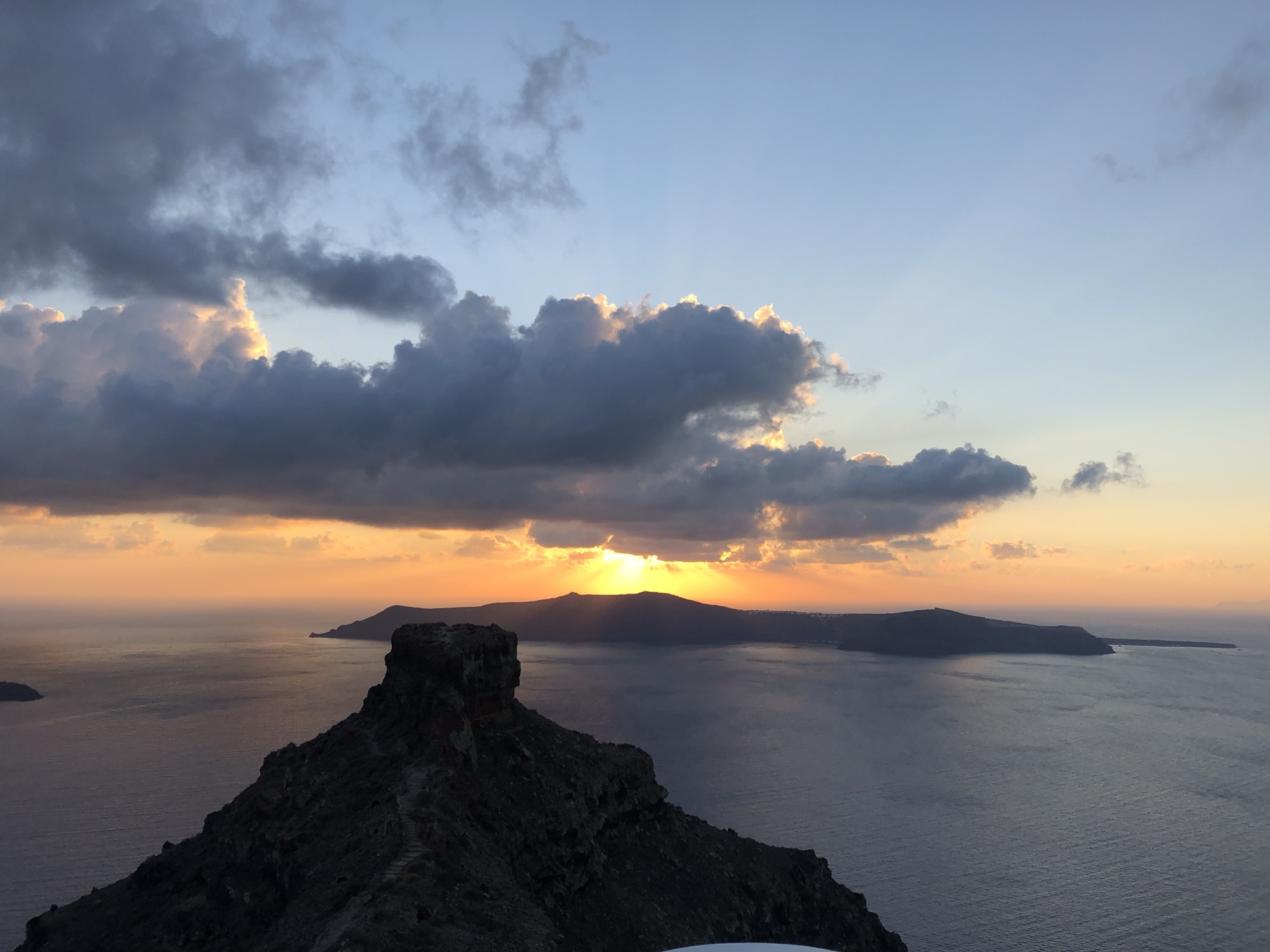 A high-angle, wide shot captures a sunset over a body of water, with the sun dipping behind a distant island and casting golden light through the clouds. In the foreground, a prominent, rocky cliffside overlooks the sea under a partly cloudy sky.