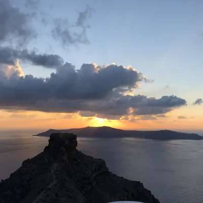 A high-angle, wide shot captures a sunset over a body of water, with the sun dipping behind a distant island and casting golden light through the clouds. In the foreground, a prominent, rocky cliffside overlooks the sea under a partly cloudy sky.