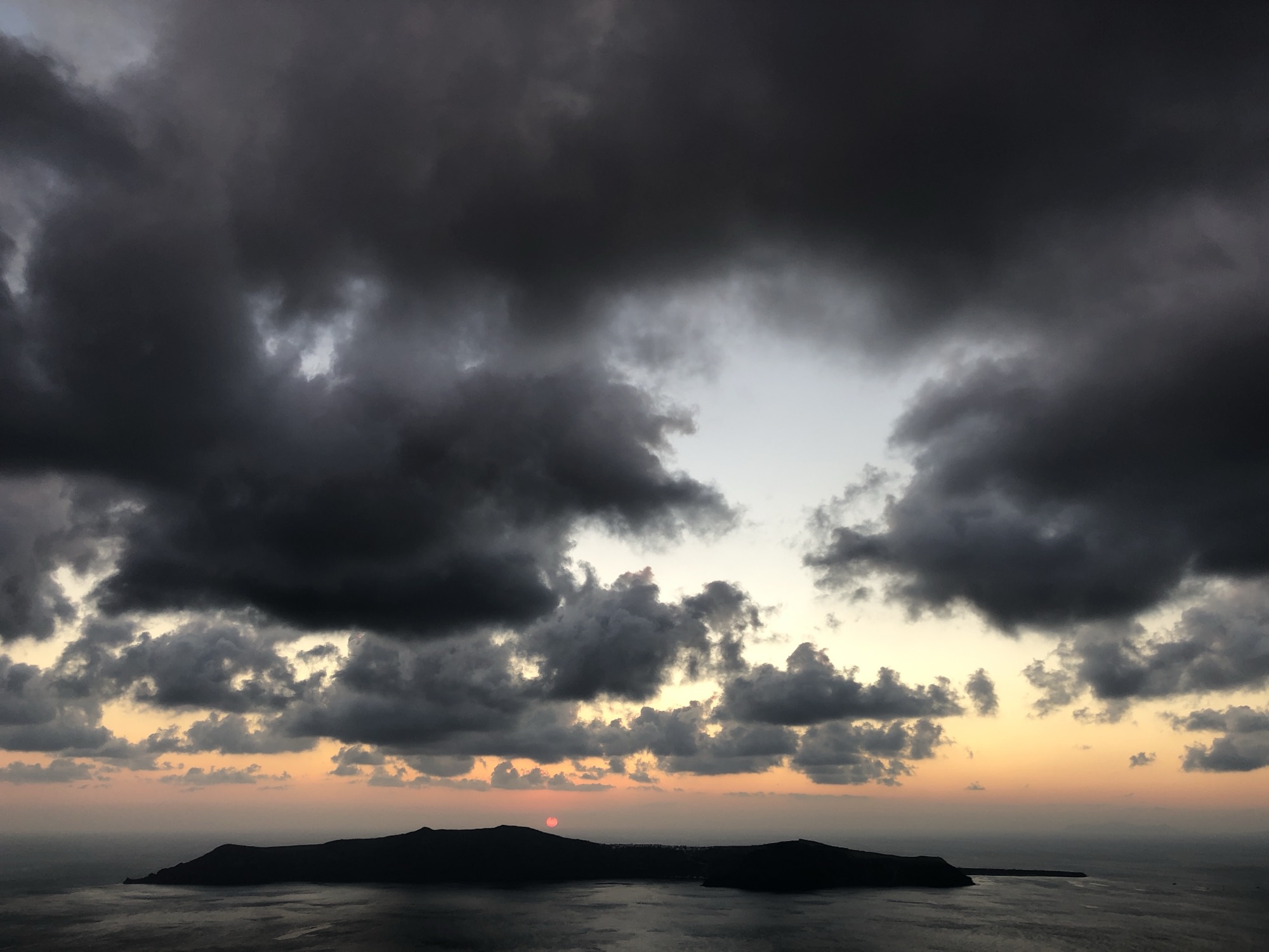 A wide shot captures a sunset over a dark, silhouetted island in the sea, with the sun dipping below the horizon under a dramatic sky filled with heavy, dark clouds. The sky transitions from a bright orange glow near the horizon to a pale blue and deep grey above.