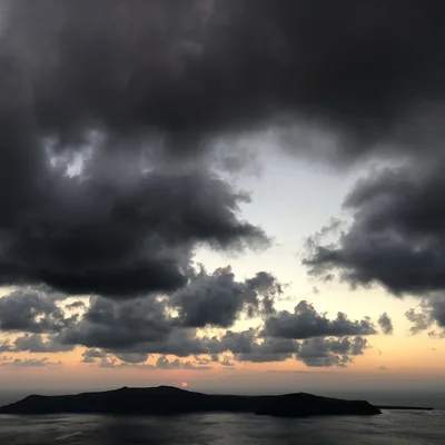 A wide shot captures a sunset over a dark, silhouetted island in the sea, with the sun dipping below the horizon under a dramatic sky filled with heavy, dark clouds. The sky transitions from a bright orange glow near the horizon to a pale blue and deep grey above.