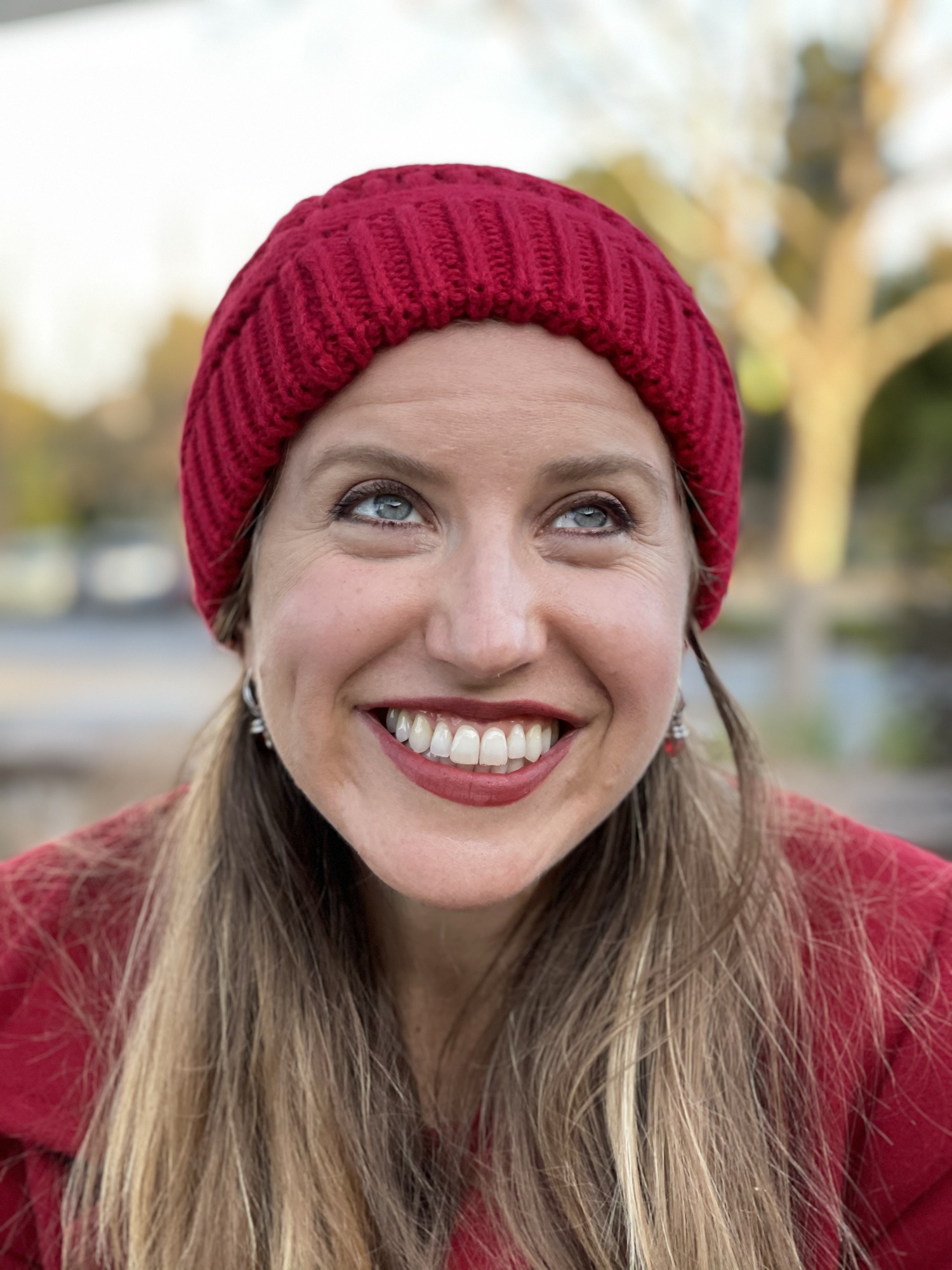 A close-up portrait of a smiling woman wearing a red knit beanie and a red coat, looking upward and to her right. She is outdoors with a softly blurred background of trees and a street.